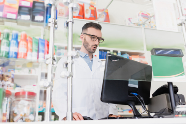 farmacista sorpreso in camice bianco e occhiali guardando monitor del computer in farmacia - Foto, immagini