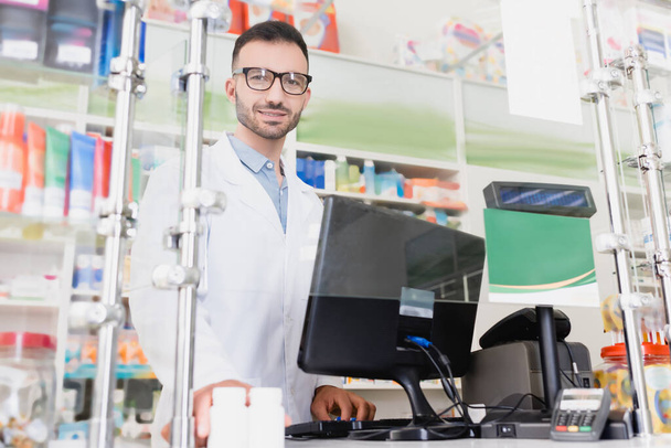 бородатый фармацевт в белом халате и очках улыбается возле монитора компьютера в аптеке - Фото, изображение