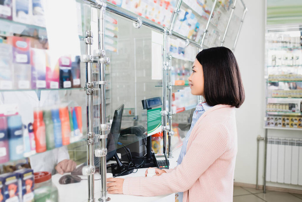 Seitenansicht eines asiatischen Kunden, der Flaschen mit Pillen in der Nähe eines Verkäufers in der Drogerie hält  - Foto, Bild
