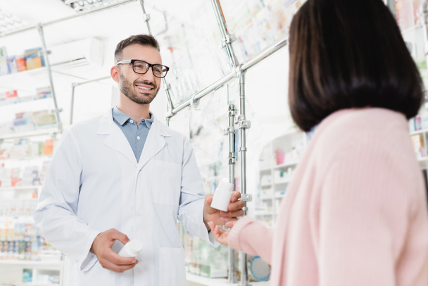 alegre farmacéutico en gafas y abrigo blanco sosteniendo botellas con pastillas cerca del cliente en primer plano borrosa - Foto, Imagen