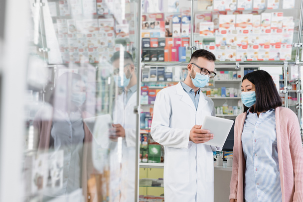 farmacista in maschera medica in possesso di tablet digitale vicino alla donna asiatica in farmacia - Foto, immagini