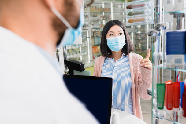 asiático cliente en médico máscara apuntando con dedo cerca farmacéutico en borrosa primer plano - Foto, imagen