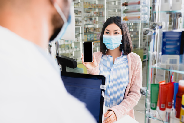 mujer asiática en máscara médica mostrando teléfono inteligente con pantalla en blanco a farmacéutico en primer plano borrosa - Foto, imagen