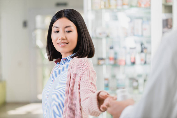 веселая азиатка, принимающая бутылку с таблетками рядом с фармацевтом на размытом переднем плане - Фото, изображение