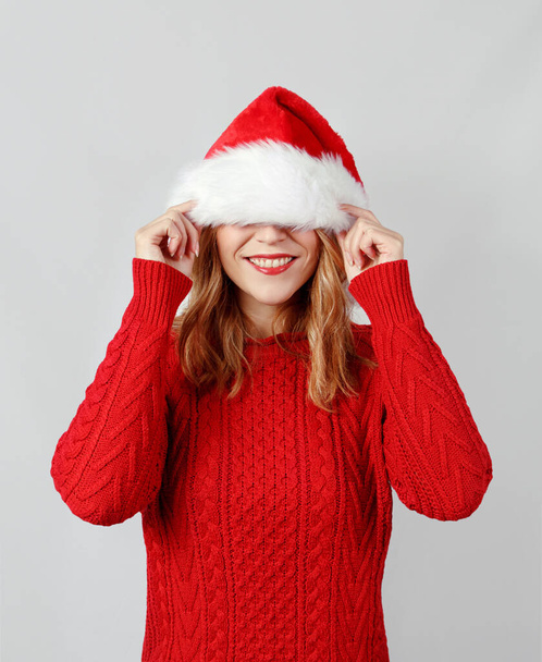 Mladá žena zakrývá oči, zatímco dotýká Santa klobouk izolované na šedé - Fotografie, Obrázek