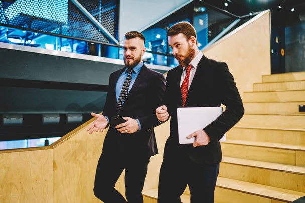 Confident male owners of company walking at office stairs have consultancy conversation holding laptop computer, formally dressed colleagues discussing financial exchange planning productive strategy - Photo, image