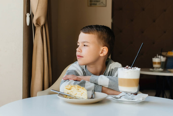 Um menino bonito está sentado em um café e olhando para um bolo e cacau close-up. Dieta e nutrição adequada - Foto, Imagem