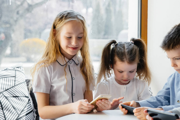 Children sit at a table in a cafe and play mobile phones together. Modern entertainment - Фото, зображення