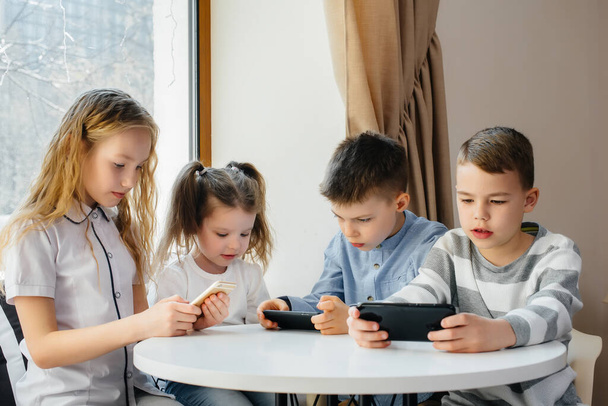 Children sit at a table in a cafe and play mobile phones together. Modern entertainment - 写真・画像