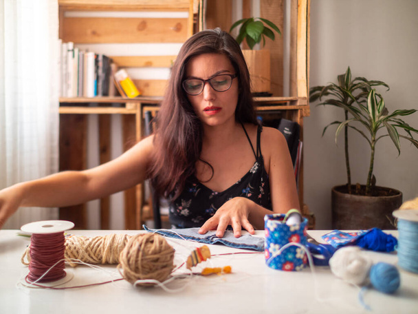 A Young Attractive Hispanic Woman in Black Dress is on a Table Making Crafts with Cloth and Threads - 写真・画像