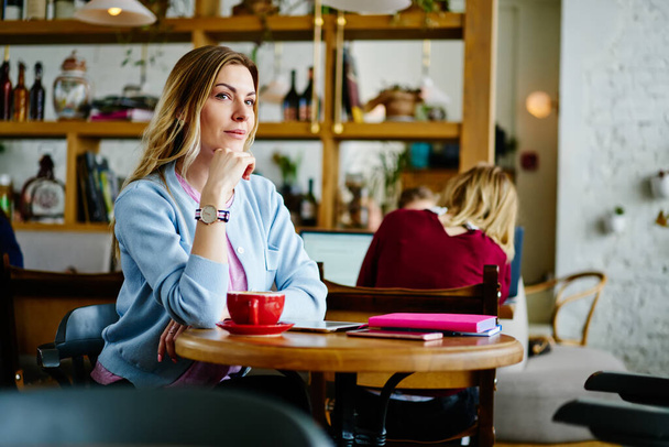 Pensive female in casual clothes sitting at wooden table and drinking cup of hot coffee while resting after workday in cozy cafe - Zdjęcie, obraz