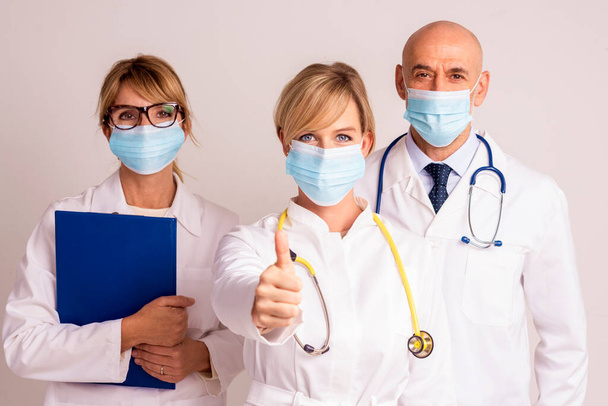Shot of medical team wearing face masks and standing together at isolated background while female doctor giving thumbs up. - Фото, изображение