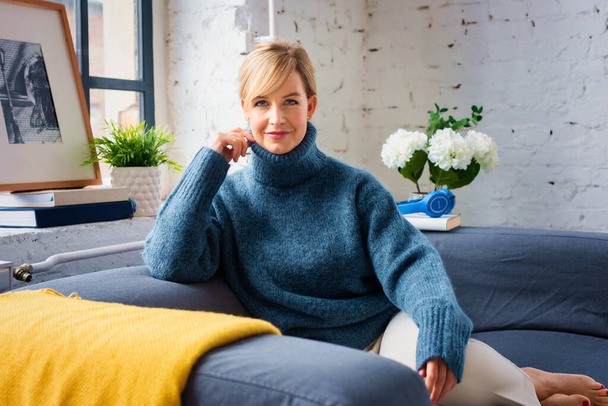 Portrait shot of beautiful blond haired woman wearing turtleneck sweater while relaxing on sofa at home.  - Foto, afbeelding
