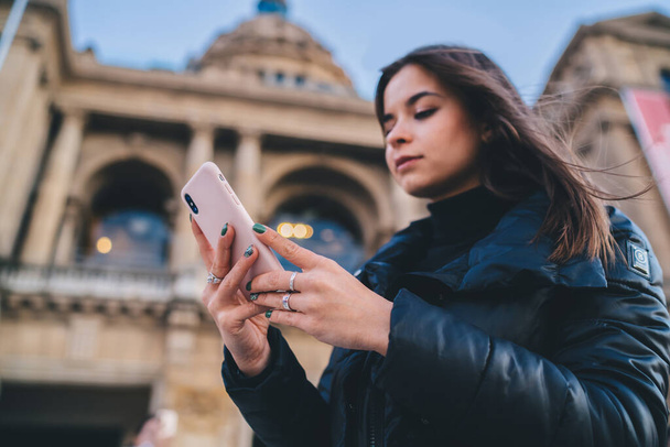 Low angle of young calm woman tourist in warm clothes browsing smartphone and checking orientation while sightseeing and exploring city against blurred facade of building - Fotoğraf, Görsel