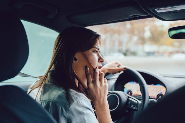 Side view of concentrated young female driver in casual clothes sitting in car at steering wheel in city and talking on smartphone - Fotoğraf, Görsel