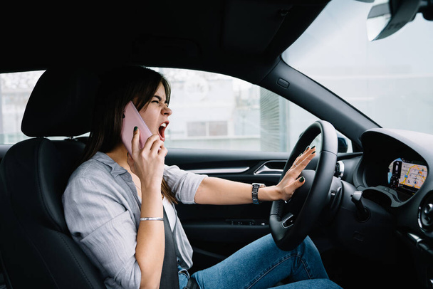 Vista lateral de la joven enojada con ropa casual sentada en el coche y gritando mientras habla en el teléfono inteligente y mirando la carretera - Foto, imagen