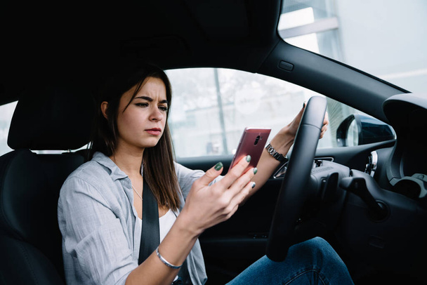 Misunderstand young woman in casual clothes sitting in car and browsing mobile phone while looking at screen on city road - Foto, imagen