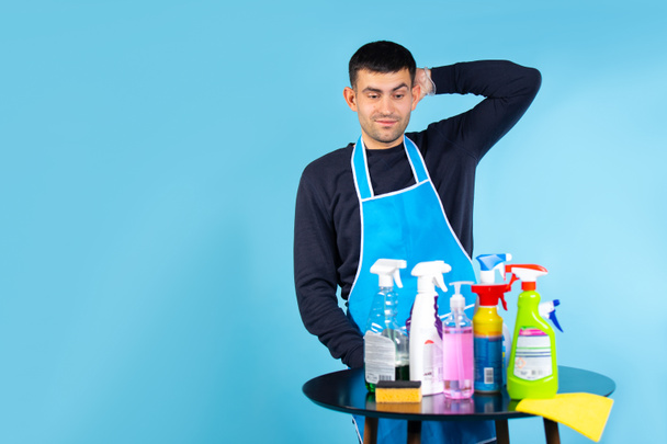 Gender stereotypes. The man holds his head and looks at the set of cleaning products. Photo on a blue background with empty side space. - Фото, зображення