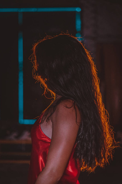 Een ondiepe focus van een vrouw met lang bruin haar in een rode jurk onder het licht in een donkere kamer - Foto, afbeelding