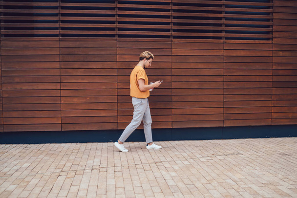 Side view of young modern concentrated guy messaging smartphone attentively during walking against wooden town wall with coffee to go - Foto, immagini