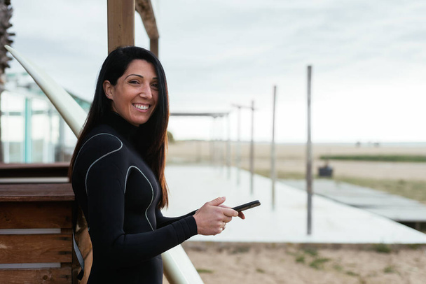 A Caucasian woman wearing a surfing suit holding a phone and happily smiling - Foto, imagen