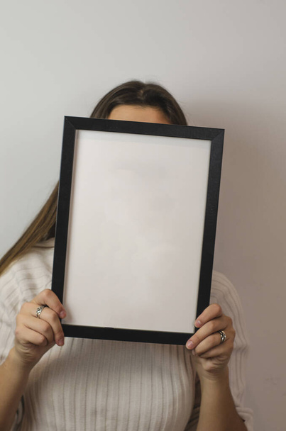 Um tiro vertical de uma mulher segurando um papel branco claro sinal branco com uma moldura preta cobrindo seu rosto - Foto, Imagem