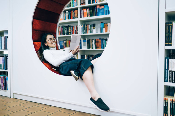 Feliz mujer étnica pacífica en anteojos y ropa casual escalofriante en elegante banco suave en la librería moderna mientras lee libro emocionante - Foto, imagen