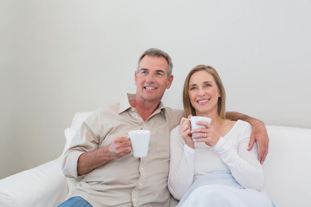 Loving couple with coffee cups looking away in living room - Foto, Bild