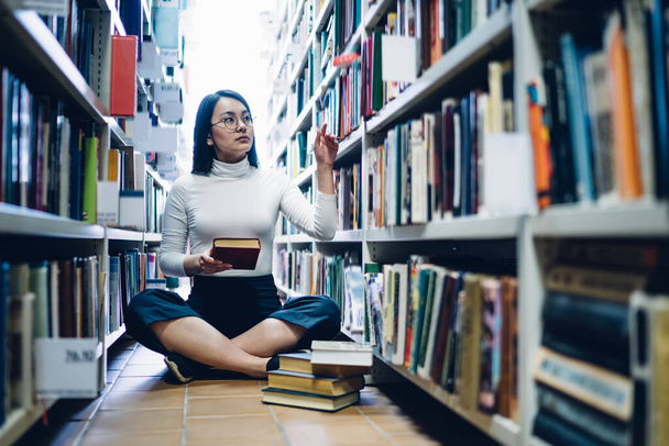Pretty serious young Asian female choosing books from shelf in library while sitting on floor with crossed legs in front of stack of books   - Foto, imagen