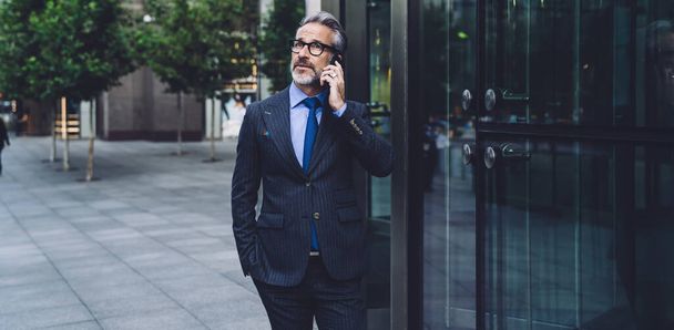 Attractive fashionable businessman with beard in stylish dark business suit talking on phone near office building in city street and looking up in New York - Photo, Image