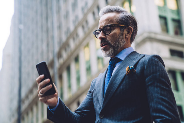 From below side view of middle aged businessman in glasses and stylish dark business suit with smartphone on background of high building in New York - Foto, Imagem