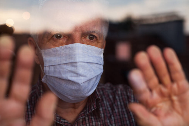 Anciano caucásico con máscara facial protectora hecha a mano, en un hogar de ancianos, mirando fuera de la ventana, tristeza, estrés y esperanza en sus ojos, auto aislamiento debido a la pandemia global de Coronavirus COVID-19 - Foto, imagen