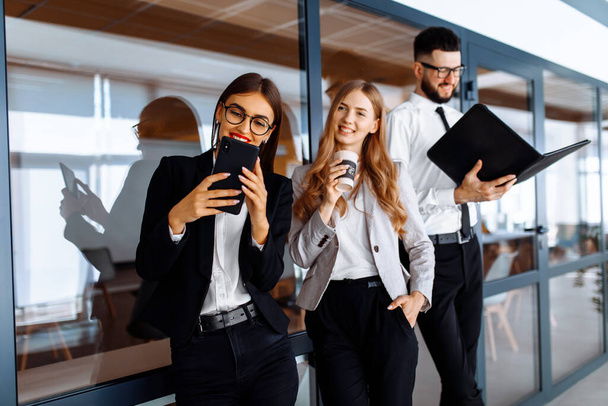 Group of attractive business people standing and communicating together, holding cups, in a modern office. Coffee break. - 写真・画像