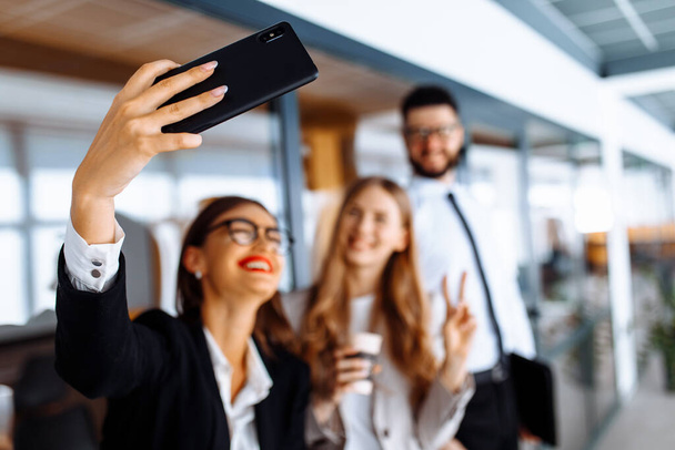 Group of attractive business people standing and chatting together holding coffee cups and taking selfie on mobile phone office - Foto, afbeelding