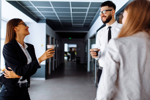 Gruppe attraktiver Geschäftsleute, die in einem modernen Büro stehen und miteinander kommunizieren und Tassen in der Hand halten - Foto, Bild