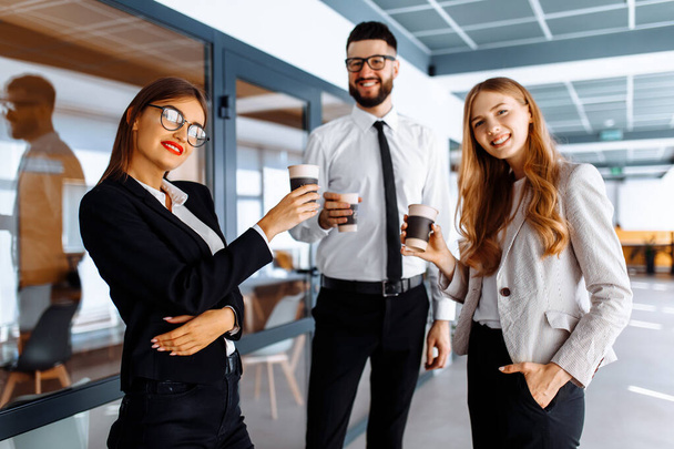 Młodzi, szczęśliwi biznesmeni stojący i rozmawiający ze sobą, trzymający kubki w nowoczesnym biurze. Przerwa na kawę. - Zdjęcie, obraz