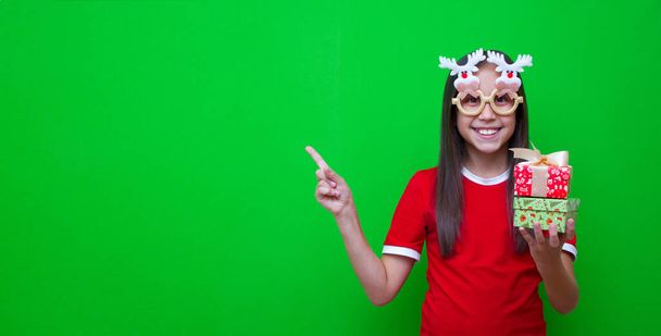 Ein Mädchen mit dunklen Haaren, rotem T-Shirt und lustiger Brille in der Hand hält Geschenke für Weihnachten und Neujahr in der Hand und zeigt mit dem Finger auf die Werbestelle..  - Foto, Bild
