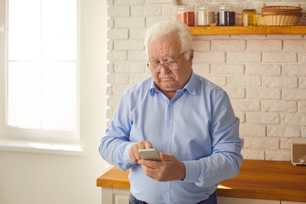 Serious senior man in glasses standing in the kitchen and using modern mobile phone - Photo, Image