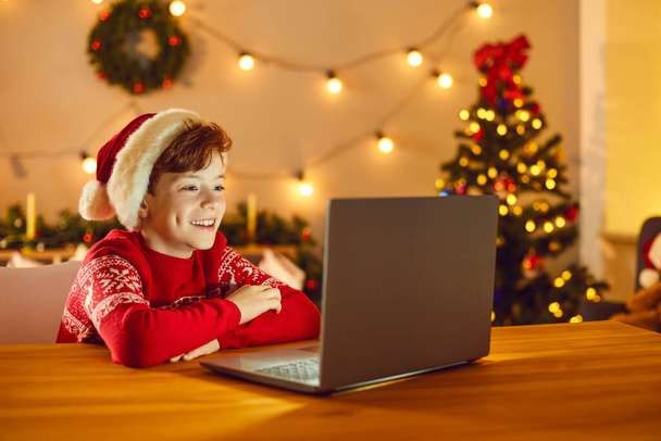 Happy boy in festive red Santa hat and sweater sitting and watching Christmas movie online on laptop - Photo, image