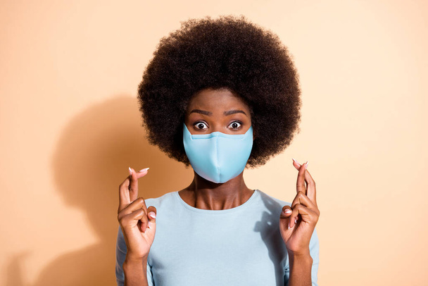 Portrait of anxious african woman holding fingers crossed wear medical respiratory mask isolated beige color background - Foto, imagen
