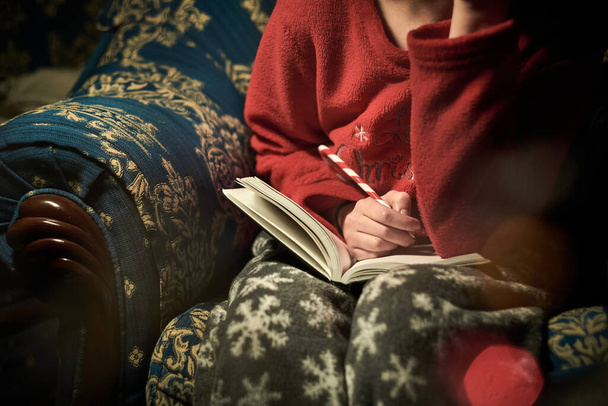 niña en pijama de Navidad rojo y gris con copos de nieve en el sofá. adolescente escribiendo en cuaderno con lápiz de Navidad. lápiz en forma de palo de caramelo y mensaje escrito - Foto, imagen
