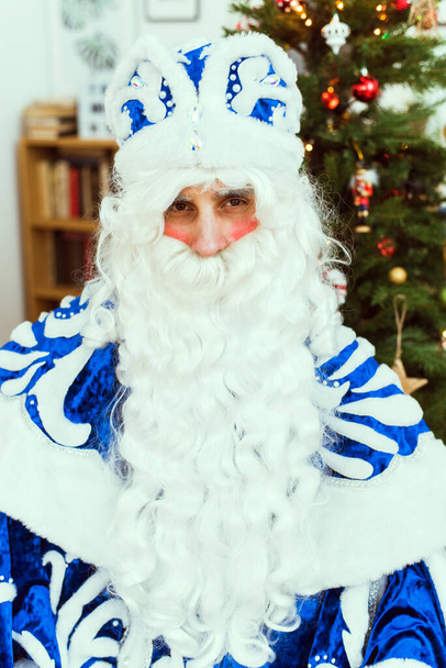 Санта-Клаус в синем шубе сидит в теплой комнате и смотрит в камеру. Концепция Нового года и Рождества - Фото, изображение