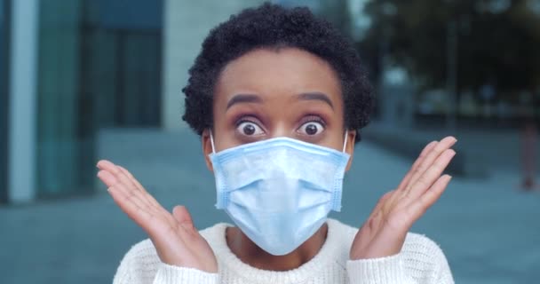 Gros plan de la femme surprise d'origine africaine portant un masque médical sur le visage féminin gesticule activement avec ses mains est choqué étonnement dit wow émotion de l'horreur se tient à l'extérieur près du bâtiment - Séquence, vidéo