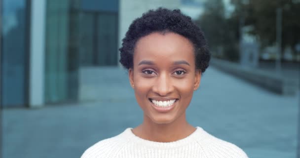 Portrait de belle heureuse sortie élégante afro-américaine étudiante fille modèle féminin toothy souriant à la caméra regardant avec confiance portant des vêtements blancs posant sur fond urbain, gros plan - Séquence, vidéo