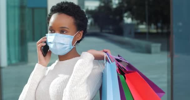 Retrato afroamericano mujer shopper shopaholic adolescente usa máscara médica en su cara habla por llamadas de teléfono móvil taxi para almacenar tiene bolsas de compras brillantes en sus manos, venta durante la cuarentena - Metraje, vídeo