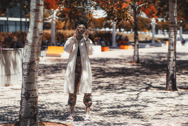 A stylish young black female in a white trench and camouflage pants, with curly Afro hair is adjusting her sunglasses while standing on a paving-stone in the shadow of autumn poplar trees in a park - Zdjęcie, obraz