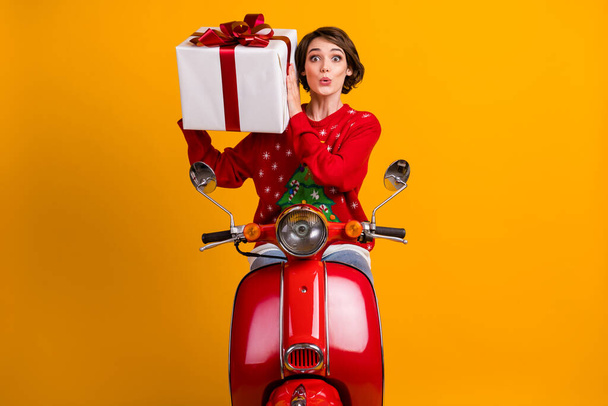 Photo of astonished girl in christmas tree ornament sweater pullover shake gift box drive red scooter x-mas eve noel party isolated over bright shine color background - 写真・画像