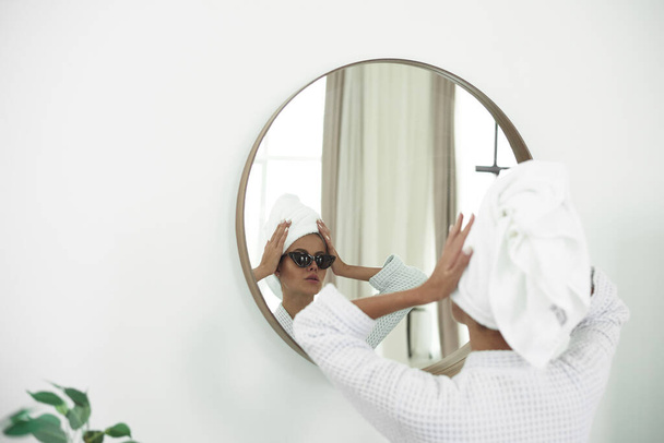 Молода жінка в халаті, з білим рушником на голові і чорними сонцезахисними окулярами дивиться у дзеркало ванної кімнати
 - Фото, зображення