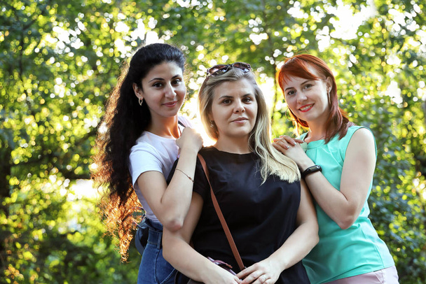 Three young smiling women friends look and pose for the camera against the backdrop of foliage of trees on summer sunny day. Friendship and ethnic diversity concept - Photo, Image