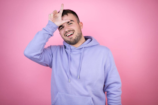 Молодий красивий чоловік у повсякденному светрі над ізольованим рожевим тлом робить сміх людям з пальцями на лобі робить жест невдахою, глузуючи і ображаючи
. - Фото, зображення